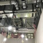 Exposed HVAC ductwork, pipes, and electrical wiring on a ceiling under construction by Sierra Trading Commercial HVAC Spokane Valley, with unfinished walls and insulation visible. Bright work lights hang from the ceiling.