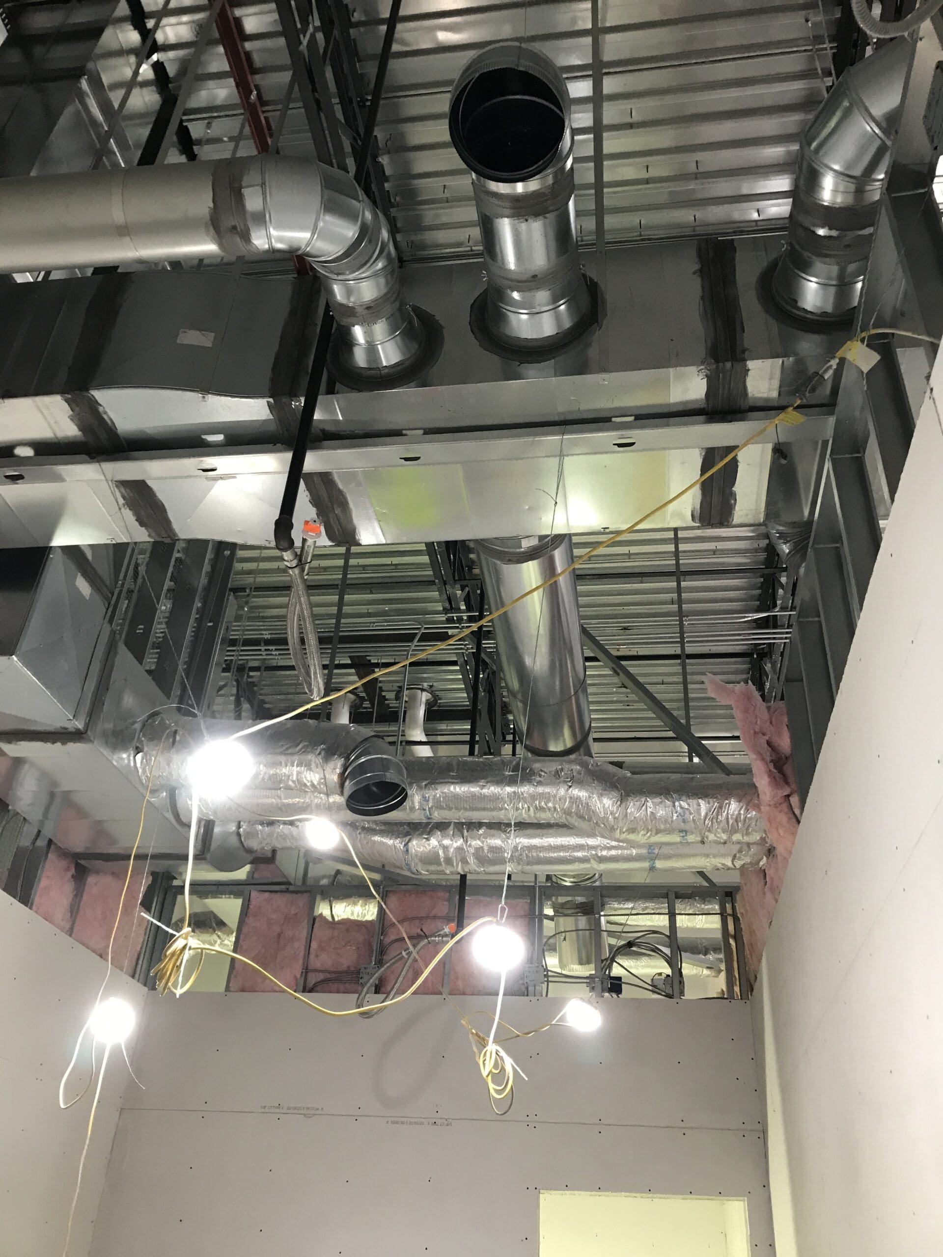 Exposed HVAC ductwork, pipes, and electrical wiring on a ceiling under construction by Sierra Trading Commercial HVAC Spokane Valley, with unfinished walls and insulation visible. Bright work lights hang from the ceiling.