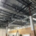 Exposed ductwork and metal pipes on the ceiling of a large unfinished commercial building, featuring ladders and construction materials on the floor—a typical scene for Sierra Trading Commercial HVAC Spokane Valley projects.