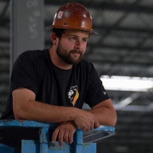A man wearing a brown hard hat and black t-shirt stands leaning on a blue railing inside an industrial building, embodying the spirit of HVAC innovation in commercial HVAC solutions.