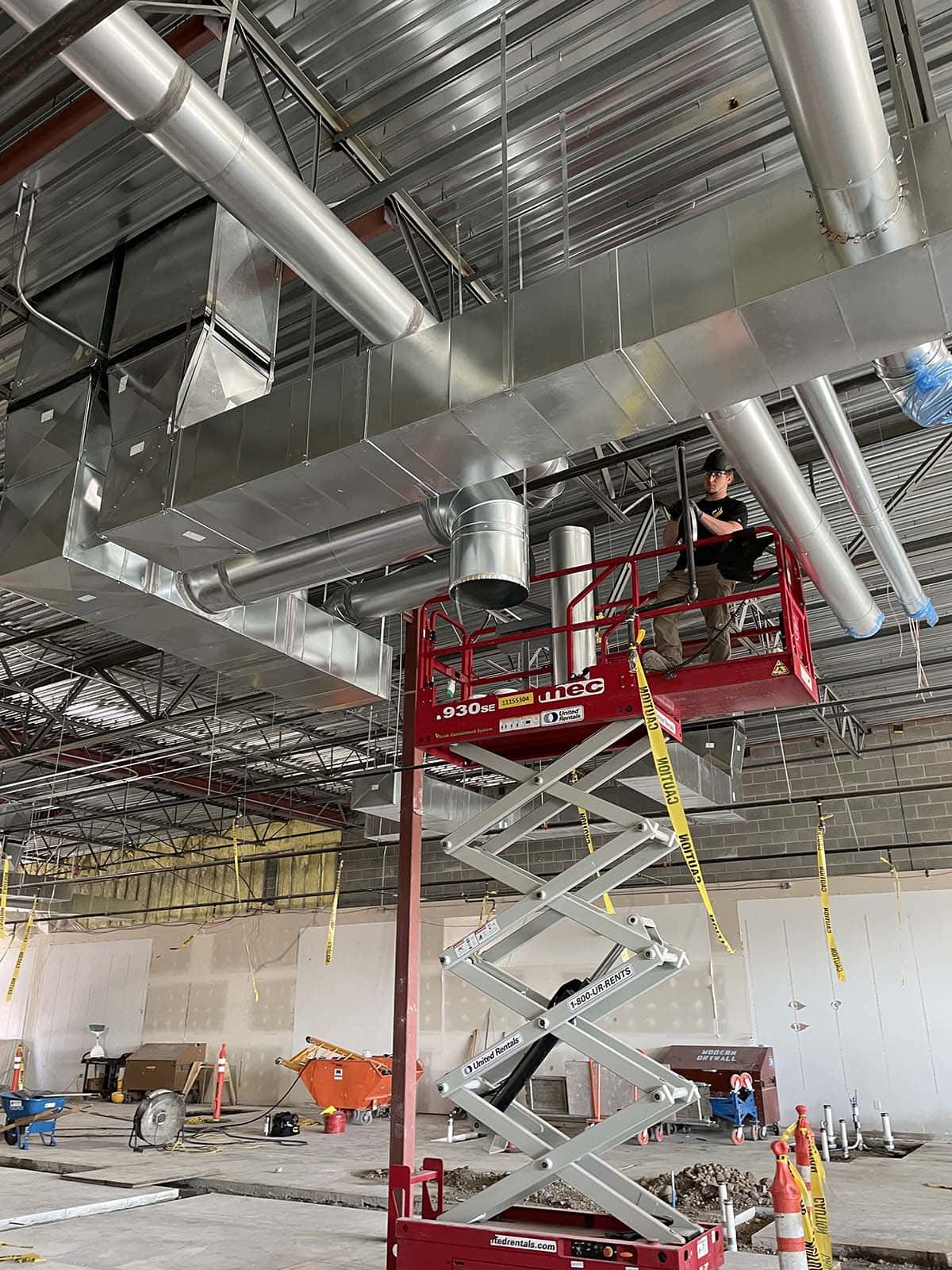 A worker stands on a scissor lift installing or inspecting HVAC ductwork on the ceiling inside a large building under construction.