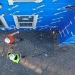 Two construction workers in hard hats stand on gravel next to a building covered in blue weatherproofing material—part of the University of Idaho HVAC project—viewed from above.