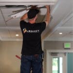 A worker in a "Legacy" shirt installs or repairs wiring in a ceiling panel of a commercial building hallway for the University of Idaho HVAC project.