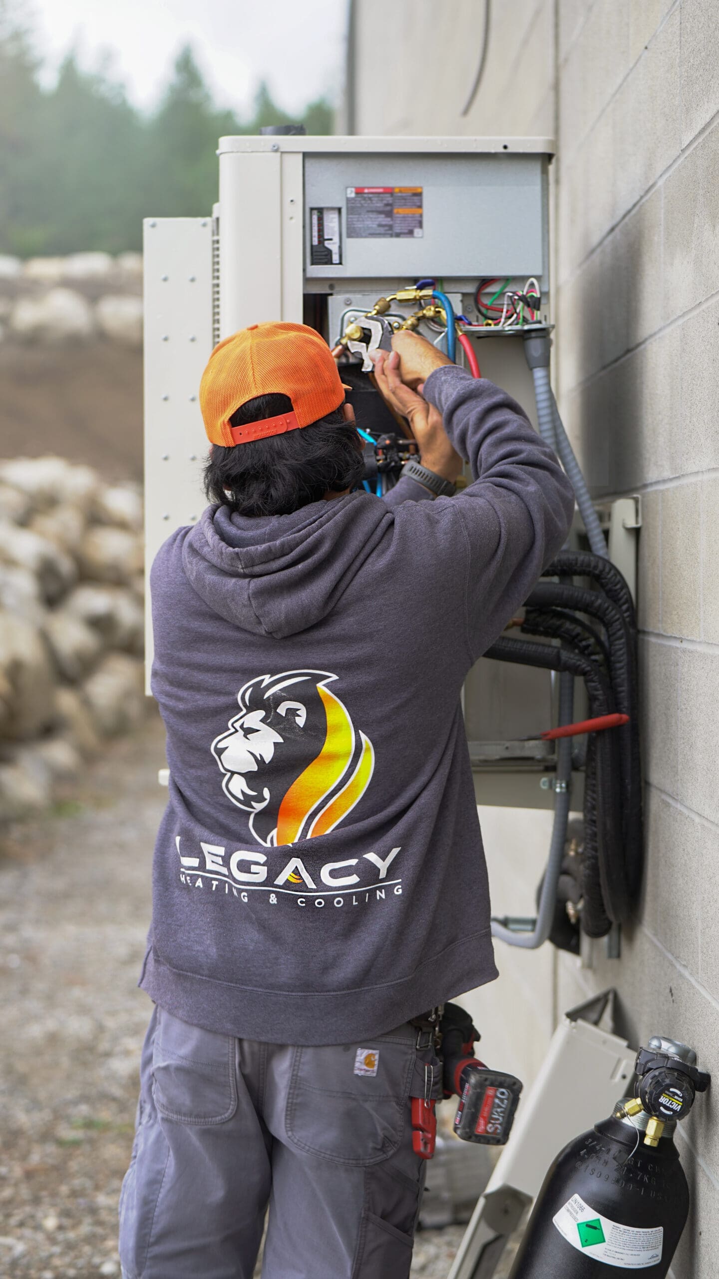 A technician wearing an orange cap and "Legacy Heating & Cooling" sweatshirt works on an outdoor electrical panel, performing spring HVAC prep on a building wall.